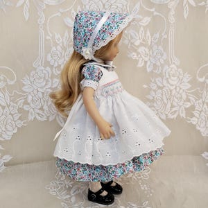Puede incluir: Una muñeca vestida con un vestido blanco con detalles de ojal y un dobladillo con estampado floral. La muñeca tiene el pelo largo y rubio y lleva un gorro a juego con una cinta blanca. Los zapatos Mary Jane negros completan el conjunto.