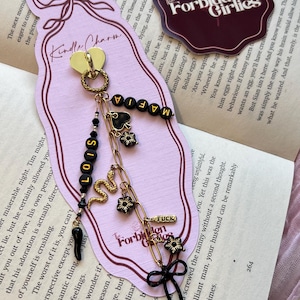 Puede incluir: Un charm decorativo para marcapáginas con un corazón dorado, cuentas negras que deletrean "LOIS" y "MAFIA", y varios dijes. El charm incluye una serpiente, una pica y un lazo, con la palabra "FUCK" en una pequeña etiqueta. El fondo es un libro.