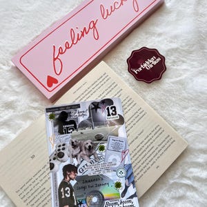 Puede incluir: Una caja rectangular rosa con la frase "feeling lucky" en escritura roja y corazones. También se ve una pegatina con "Forbidden Girlies" y un collage de imágenes y texto.