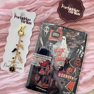 May include: A Halloween-themed phone case featuring a collage of spooky imagery, including ghosts, skeletons, and pumpkins. A charm with a crescent moon, beads, and a skeleton is also present. The text "Trick or Treat" is visible.
