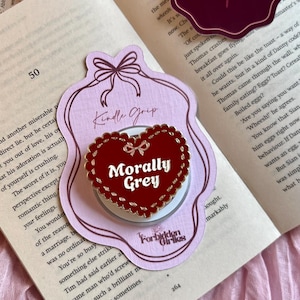 Könnte beinhalten: Ein herzförmiger Kindle Grip mit dem Text "Morally Grey" in Weiß, umgeben von einem roten Rand. Der Griff befindet sich auf einem hellrosa Anhänger mit einer Schleife und den Worten "Kindle Grip". Der Hintergrund umfasst ein offenes Buch und ein "Forbidden Girlies"-Logo.