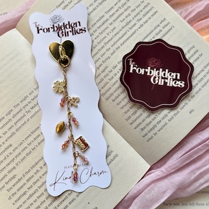 May include: Gold-coloured charm featuring a heart, clover, football, and book charms, with pink beads. The charm is on a white card with "The Forbidden Girlies" and "Kindle Charm" text.