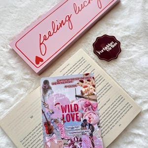 Può includere: Una scatola rosa con le parole "feeling lucky" in corsivo, accompagnata da due cuori. Un adesivo con la scritta "Forbidden Girlies" è accanto a un collage con le parole "Wild Love". Gli oggetti sono su una superficie bianca e soffice.