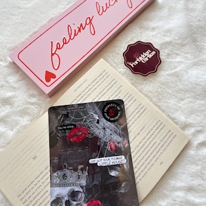 以下が含まれることがあります： 「feeling lucky」と赤い文字で書かれたピンク色の長方形の箱、"Forbidden Girlies"の文字が入ったステッカー、そして骸骨、バラ、テキストが描かれたカード。これらのアイテムは開いた本の上に置かれています。