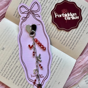 Puede incluir: Pendiente decorativo con un corazón blanco y negro, cuentas rojas y la palabra "SPICY" en letras rojas. El pendiente también presenta un pequeño dije de pistola y un dije de carta de juego. Texto "Forbidden Girlies" en una tarjeta rosa.