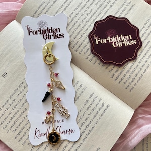 Könnte beinhalten: Ein handgefertigter Kindle-Charm mit einem goldfarbenen Halbmond, einem schwarzen und goldenen kreisförmigen Charm und verschiedenen Charms. Der Charm enthält ein Skelett, einen Flügel, eine Chilischote und rote Perlen. Der Text "The Forbidden Girlies" ist sichtbar.