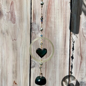 May include: A hanging decorative piece featuring a dark green heart-shaped stone within a clear circular frame. The piece is adorned with various beads and a dark green, bell-shaped stone at the bottom, suspended by a gold chain.