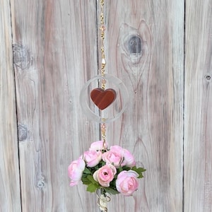 May include: A decorative hanging ornament featuring a red heart within a clear circle, suspended by a gold chain with pink beads. Below, a cluster of pink roses with green leaves adds a floral touch.
