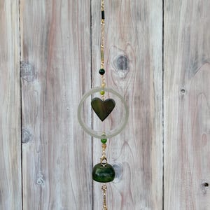 May include: A hanging decorative piece featuring a dark green heart pendant, a circular ring, and a rounded green stone. The piece is strung with gold-colored chain and beads, with a wooden background.