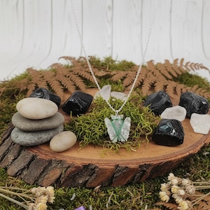 May include: A silver necklace featuring a clear, arrowhead-shaped pendant secured with green wire. The necklace is displayed on a wooden slice, surrounded by moss, stones, and crystals. Dried flowers and a purple ribbon complete the natural theme.