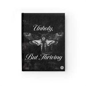 Puede incluir: Un diario negro con las palabras "Unholy, But Thriving" en escritura blanca. La portada presenta una ilustración detallada de una polilla con un patrón de calavera en su cuerpo y alas. El fondo tiene una apariencia texturizada y desgastada.