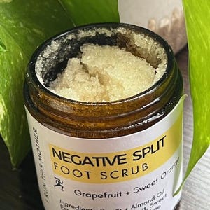 May include: A jar of foot scrub with a yellow and white label. The label reads "NEGATIVE SPLIT FOOT SCRUB" with the ingredients listed below. The scrub is light brown and granular. The jar is surrounded by green leaves.