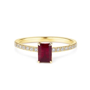 May include: A gold ring featuring a rectangular, deep red gemstone. The band is embellished with small, clear gemstones. The ring is displayed against a white backdrop, accentuating its design and details.