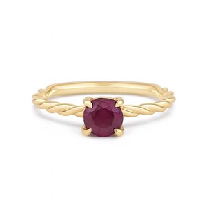 May include: A gold ring with a twisted band. It features a round, deep red ruby gemstone held in place by four prongs. The ring is set against a white background.