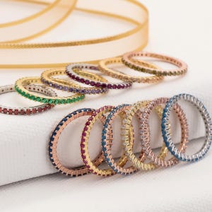 May include: A collection of colourful eternity rings with gemstones in shades of red, green, purple, and blue. The rings are displayed on a white surface, with a gold ribbon in the background.