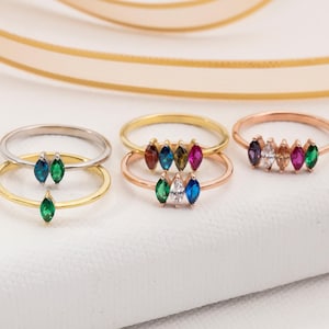 May include: A collection of gemstone rings in gold, silver, and rose gold. Each ring features marquise-cut gemstones in various colors, including green, blue, pink, and white. The rings are displayed on a white surface with a gold ribbon in the background.
