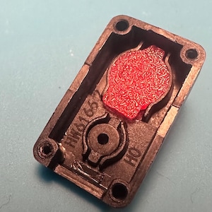 May include: A black rectangular plastic component with a red textured insert. The component has four holes in the corners and the text "HW6165" and "HO" printed on it. The red insert has a unique pattern.