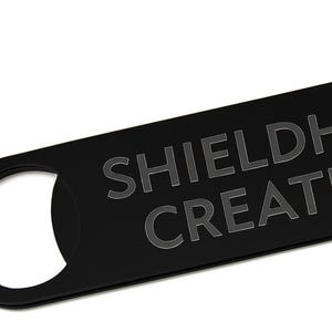 May include: Black bottle opener with the text "SHIELDHAVEN CREATIONS" in a sans-serif font. The opener has two circular cutouts, one at each end. The background is white.