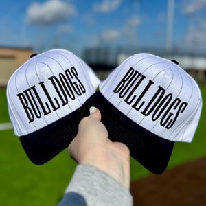 May include: Two white baseball caps with black brims and the word "BULLDOGS" in black script. The caps have a pinstripe design and are held up against a blue sky with clouds and a green field.