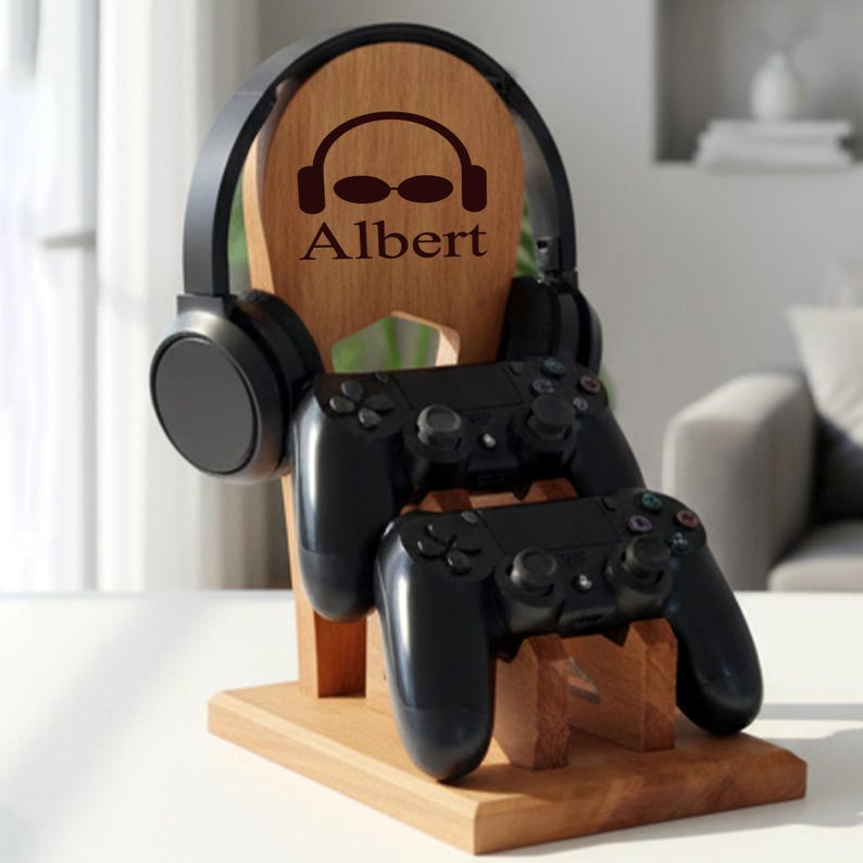 K&ouml;nnte beinhalten: Holz-Kopfh&ouml;rer- und Controller-St&auml;nder mit dem eingravierten Namen "Albert". Der St&auml;nder h&auml;lt ein Paar schwarze Kopfh&ouml;rer und zwei schwarze Gamecontroller. Der St&auml;nder ist aus hellbraunem Holz gefertigt.