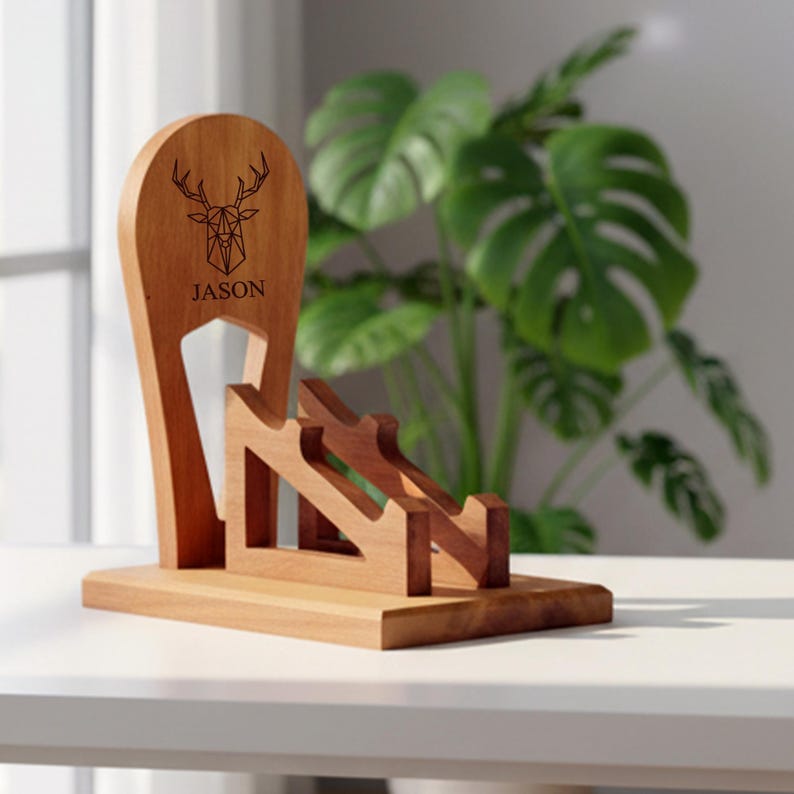 K&ouml;nnte beinhalten: Ein Handy-St&auml;nder aus Holz mit einem Hirschkopf-Design und dem eingravierten Namen "JASON". Der St&auml;nder ist aus hellbraunem Holz gefertigt und hat ein geometrisches Hirschkopf-Design. Der St&auml;nder ist f&uuml;r mehrere Telefone konzipiert.