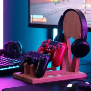 K&ouml;nnte beinhalten: Ein Holzst&auml;nder f&uuml;r Gaming-Headsets und Controller. Der St&auml;nder h&auml;lt ein schwarzes Headset und zwei Gamecontroller, einen roten und einen schwarzen. Der St&auml;nder ist aus hellem Holz gefertigt und steht auf einem Schreibtisch.
