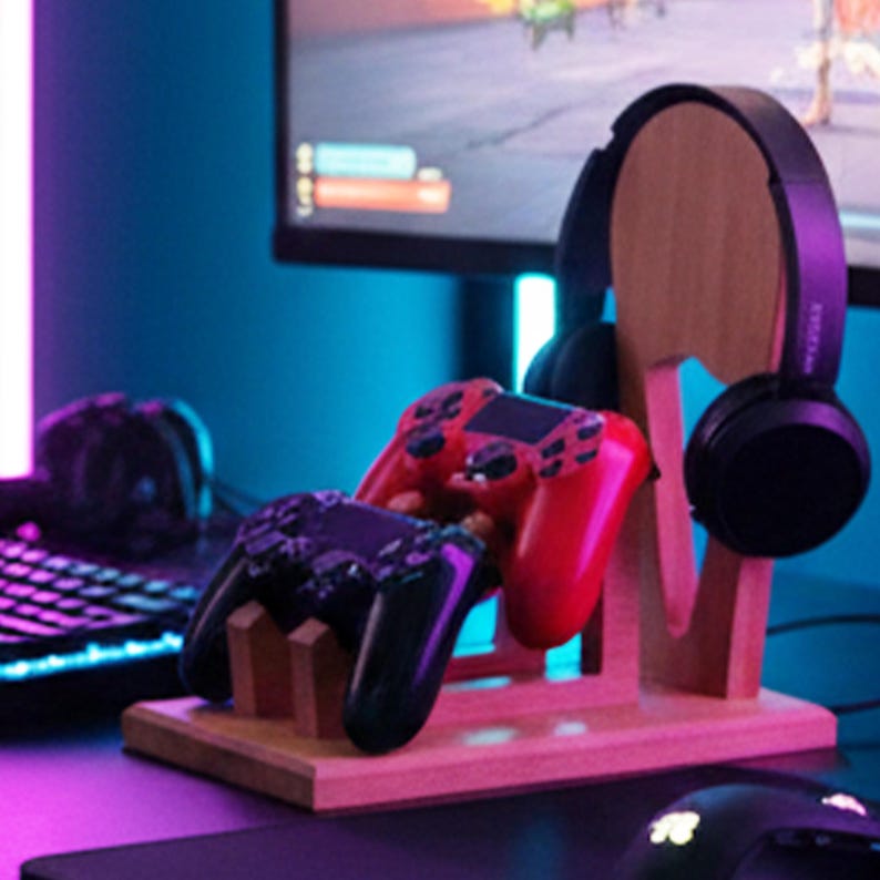 K&ouml;nnte beinhalten: Ein Holzst&auml;nder f&uuml;r Gaming-Headsets und Controller. Der St&auml;nder h&auml;lt ein schwarzes Headset und zwei Gamecontroller, einen roten und einen schwarzen. Der St&auml;nder ist aus hellem Holz gefertigt und steht auf einem Schreibtisch.