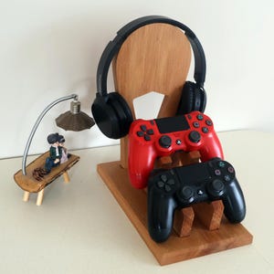 K&ouml;nnte beinhalten: Ein Gaming-St&auml;nder aus Holz mit einem schwarzen Headset und zwei Gamecontrollern, einer rot und einer schwarz. Eine kleine dekorative Lampe und eine Figur befinden sich links. Der St&auml;nder ist aus hellem Holz.