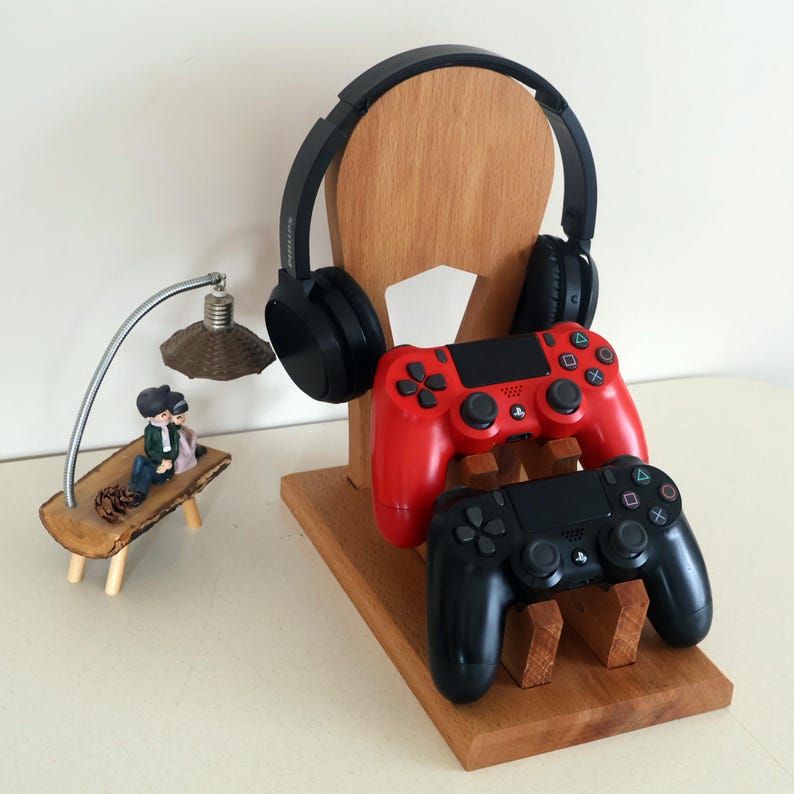 K&ouml;nnte beinhalten: Ein Gaming-St&auml;nder aus Holz mit einem schwarzen Headset und zwei Gamecontrollern, einer rot und einer schwarz. Eine kleine dekorative Lampe und eine Figur befinden sich links. Der St&auml;nder ist aus hellem Holz.