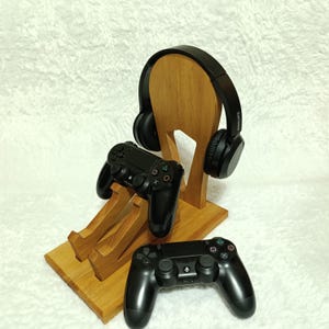 May include: A wooden gaming headset and controller stand. The stand is made of light brown wood and holds a pair of black headphones and two black game controllers. The stand has a simple, modern design.