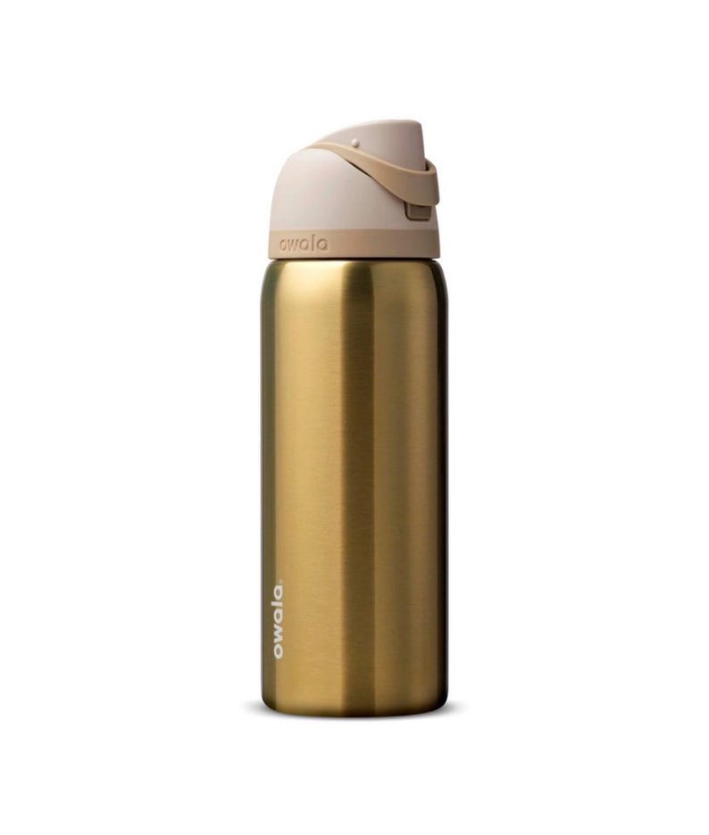 Op de afbeelding: Een goudkleurige Owala waterfles met een beige deksel en handvat. De fles heeft een slank, cilindrisch ontwerp en de merknaam staat in het wit op de zijkant. Het deksel heeft een flip-top ontwerp voor gemakkelijke toegang.