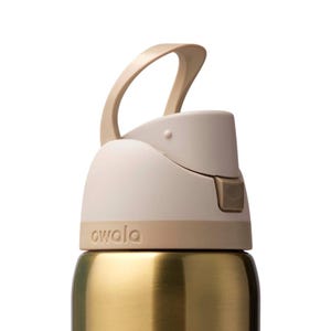 Op de afbeelding: Close-up van een goudkleurige waterfles met een beige deksel en handvat. Het deksel heeft een flip-top ontwerp en het woord "owala" staat op de basis van het deksel.
