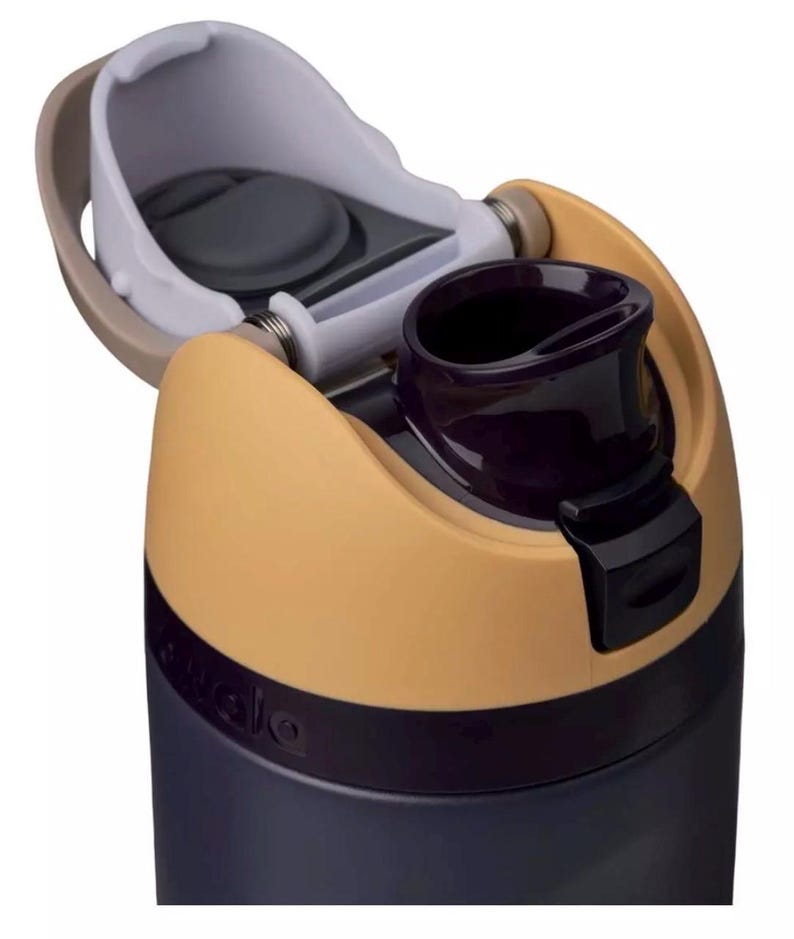 Puede incluir: Primer plano de una botella de agua con tapa abatible. La botella es principalmente azul marino con un acento beige y negro. La tapa est&aacute; abierta, revelando una boquilla para beber de color p&uacute;rpura oscuro y un interior blanco y gris.