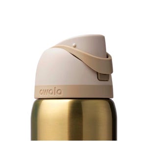 Op de afbeelding: Close-up van een goudkleurige waterfles met een beige deksel en een bruine handgreep. Het woord "owala" staat op de fles. De fles heeft een strak, modern design.