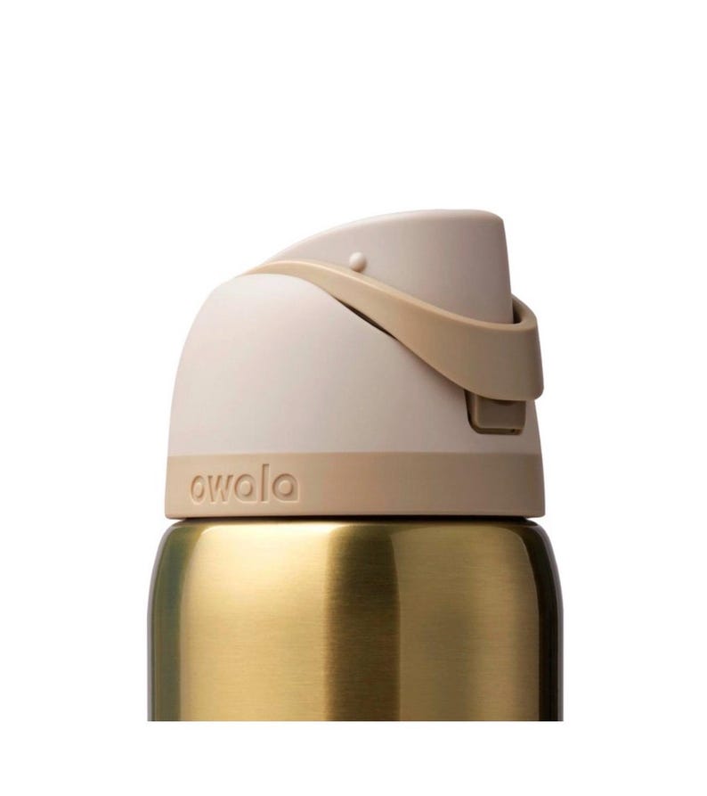 Op de afbeelding: Close-up van een goudkleurige waterfles met een beige deksel en een bruine handgreep. Het woord "owala" staat op de fles. De fles heeft een strak, modern design.