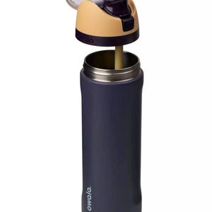 Puede incluir: Una botella de agua aislada de color azul marino con tapa abatible. La tapa es de color beige y morado, con una boquilla blanca. La botella tiene un borde de acero inoxidable y una pajita. La marca "owala" est&aacute; impresa en el lateral.