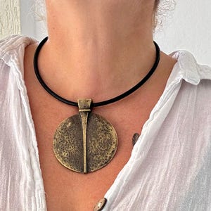 Könnte beinhalten: Eine bronzefarbene Halskette mit einem strukturierten, kreisförmigen Anhänger und einem vertikalen Liniendesign. Der Anhänger hängt an einer schwarzen Kordel. Die Halskette wird über einem weißen Hemd getragen. Der Anhänger hat einen Durchmesser von ca. 5 cm.