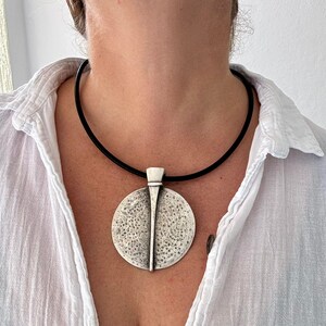 Könnte beinhalten: Silberfarbene Halskette mit einem strukturierten, kreisförmigen Anhänger und einem vertikalen Designelement. Der Anhänger hängt an einer schwarzen Kordelkette. Die Person trägt ein weißes Hemd. Modeschmuck.