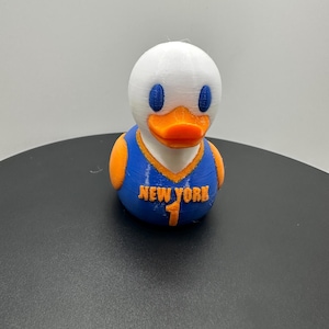 May include: A white rubber duck figurine wearing a blue basketball jersey with orange trim. The jersey features the text NEW YORK and the number 1 in orange. The duck has blue oval eyes and an orange beak, with orange wings visible on the sides.