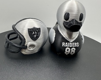 Puede incluir: Un pato de goma impreso en 3D de color plateado y negro que lleva una camiseta negra con el texto RAIDERS y el número 98, acompañado de un casco de fútbol americano plateado y negro a juego con el logotipo de los Raiders en el lateral.