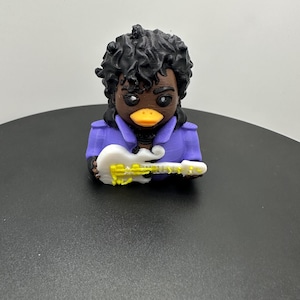 May include: A 3D-printed figurine of a duck with black curly hair, brown skin, and a yellow beak, wearing a purple jacket and holding a white and yellow guitar. The figurine is on a black surface.