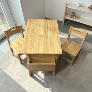 May include: A light-colored wooden children's table and two matching chairs. The table has a rectangular top and a storage compartment. The chairs have a simple design with a backrest and a cutout handle. The set is made of natural wood.