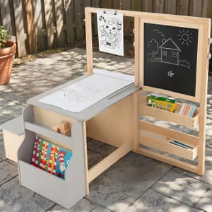 May include: A light-colored wooden art station featuring a gray tabletop with a paper roll, a black chalkboard with a house drawing, a clip holding a cat coloring page, side storage shelves for books, and two horizontal trays for art supplies.