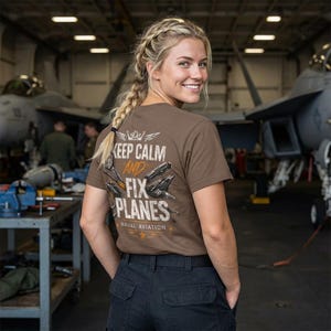 May include: A brown t-shirt with the text "KEEP CALM AND FIX PLANES NAVAL AVIATION" and an illustration of aircraft. The shirt is worn by a person in a hangar with aircraft in the background.