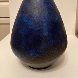 May include: A blue ceramic vase with a textured surface. The vase has a rounded base and tapers upwards to a narrower opening. The colour transitions from a dark blue at the top to a brown base.