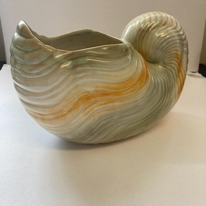 May include: A ceramic planter shaped like a nautilus shell with a glossy finish. The surface features swirling patterns in shades of pale green, cream, and warm orange, with a prominent spiral coil on one side and an open, curved top opening.