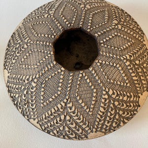 May include: A round, beige ceramic planter featuring a dark brown, recessed lace-like texture across its surface. The vessel has a hexagonal opening at the top and a small drainage hole visible at the bottom of the interior basin.