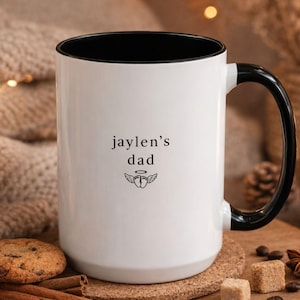 Memorial mug for angel dad, personalized baby names on white ceramic cup with black handle, lifestyle photo with warm soft lighting and minimal decor.