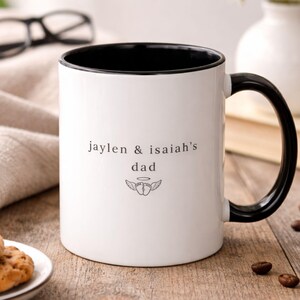 Personalized bereaved dad mug with black handle and interior, baby names and angel baby feet design on white ceramic coffee cup on rustic wood table.
