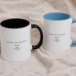 Close-up of custom baby loss dad mug showing “jaylen & isaiah’s dad” text with small angel baby symbol, white ceramic mug with black and blue rim and handle.